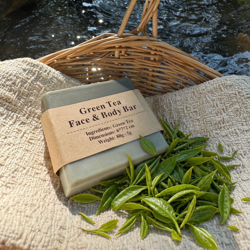 Green Tea Face & Body Bar with fresh tea leaves on textured cloth in a woven basket beside a stream