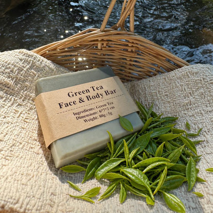 Green Tea Face & Body Bar with fresh tea leaves on textured cloth in a woven basket beside a stream