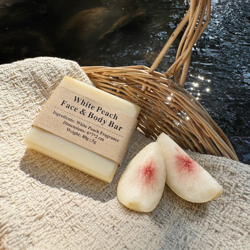 White Peach Face & Body Bar with white peach wedges on textured cloth in a woven basket beside a stream